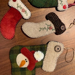 Op de afbeelding: Vier kerstsokversieringen van vilt met verschillende ontwerpen. Eén sok is rood met een groen bloemmotief, één is groen met een wit bloemmotief, één is beige met een bruine manchet en een groen bloemmotief, en één is groen en beige geruit met een sneeuwpopgezicht.