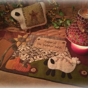 Può includere: Un tappeto patchwork marrone e verde con un girasole, un gatto e due pecore. Il tappeto ha un cartello che dice "Custode del campo di zucche".