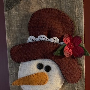 Può includere: Un pupazzo di neve di feltro con un cappello marrone, un bordo di pizzo bianco e un fiore rosso. Il pupazzo di neve ha occhi neri, un naso marrone e un viso bianco. Il pupazzo di neve è attaccato a un pezzo di legno invecchiato.