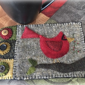 May include: A handmade felt coaster featuring a red cardinal bird with a black eye and beak, perched on a branch with green leaves and red berries. The coaster is made of gray felt with a black stitched border and has a patchwork design with green, yellow, and red felt circles.