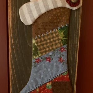 May include: A patchwork fabric stocking with a snowman face and a red flower on the top. The stocking is hanging on a rustic wooden board.