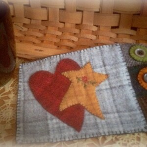 May include: A gray felt heart-shaped coaster with a yellow star and red heart applique. The coaster has a stitched border and is decorated with three felt circles with buttons in the center.