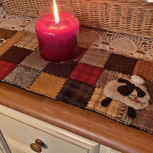 May include: A patchwork table runner with a brown, tan, red and green colour scheme. A small white sheep with black eyes is sewn onto the runner. A red candle is in a glass jar on the table.