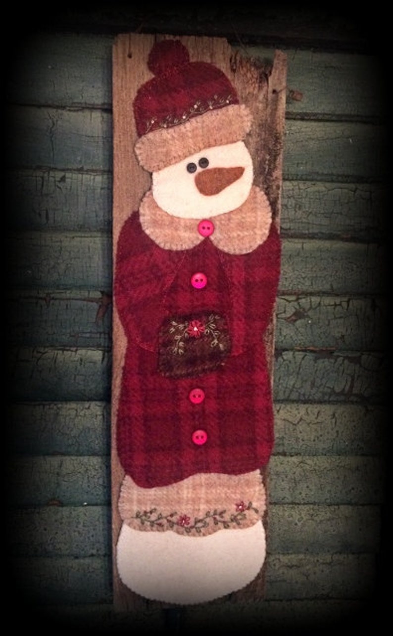 May include: A felt snowman ornament with a red coat, brown hat, and white face. The snowman is holding a small brown package and is decorated with pink buttons and green vines.
