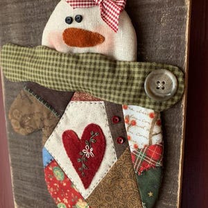 Pode incluir: Um boneco de neve de patchwork com um laço vermelho e branco, uma echarpe verde e um coração no peito. O boneco de neve está preso a um fundo de madeira rústico.