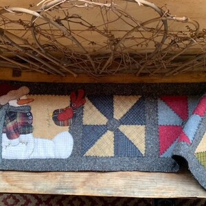 May include: A gray wool table runner with a snowman, a bird, and three patchwork quilt blocks. The blocks are in shades of blue, green, red, and tan.