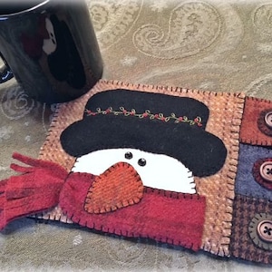 May include: A felt snowman mug rug with a black hat, red scarf, and white face. The rug has three buttons on the side.