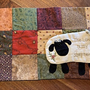 May include: A patchwork fabric item with a sheep applique and the text "The Lord is my shepherd." The item features a variety of patterned squares in shades of brown, red, and green, with a button closure.