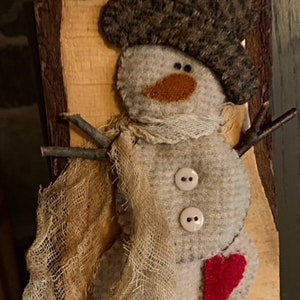 May include: A handmade felt snowman ornament with a brown felt hat, a burlap scarf, and a red felt heart. The snowman is attached to a piece of wood with a rustic finish.