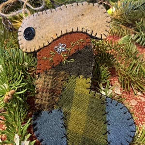 May include: A handmade felt Christmas stocking ornament with a patchwork design in shades of brown, green, blue, and orange. The stocking has a white felt flower embroidered on the top and a black button for an eye.