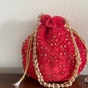 May include: A red velvet drawstring bag with gold and white beaded embellishments. The bag is decorated with a geometric pattern and has a long, beaded strap.