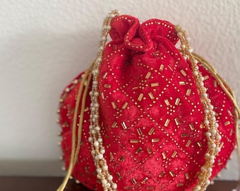 May include: A red velvet drawstring pouch featuring a diamond-patterned surface embellished with gold-toned rectangular beads and small clear crystals. The bag includes a beaded handle made of multiple strands of cream and gold-colored beads.