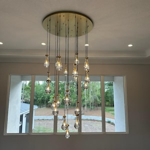 May include: A modern chandelier with a gold circular base and multiple teardrop-shaped glass bulbs hanging from it. The chandelier is suspended from the ceiling and hangs down in a cascading pattern.