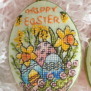 May include: A green oval-shaped fabric ornament with a yellow bead at the top. The ornament is embroidered with the words "Happy Easter" and a design of colourful Easter eggs, daffodils, and green leaves.
