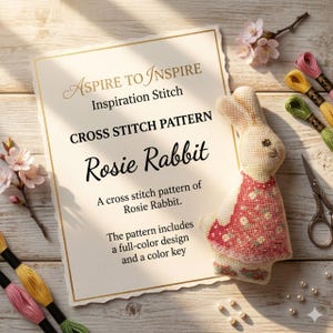 May include: A printed cross stitch pattern titled Aspire to Inspire Inspiration Stitch Rosie Rabbit, featuring a stitched rabbit figure in a red floral dress, accompanied by embroidery floss in pink, yellow, and green, and metal scissors.