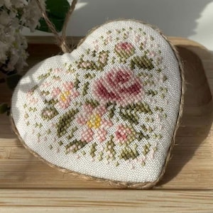 May include: A heart-shaped, cross-stitched ornament featuring a floral design in shades of pink, green, and yellow. The ornament has a natural twine border and a loop for hanging. The background is a light wooden tray.