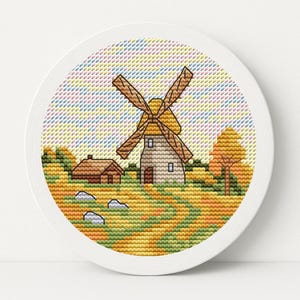 May include: A cross-stitch embroidery of a rural landscape featuring a windmill with brown blades and a small house. The scene is set in a field with autumn colors, framed in a white circular hoop.