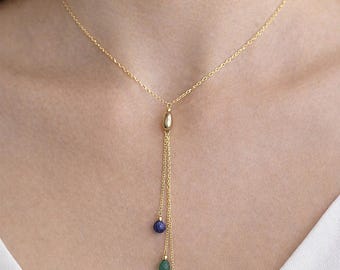 May include: A gold-colored necklace with a delicate chain and three dangling gemstones. The gemstones are blue, green, and red. The necklace is worn around the neck.