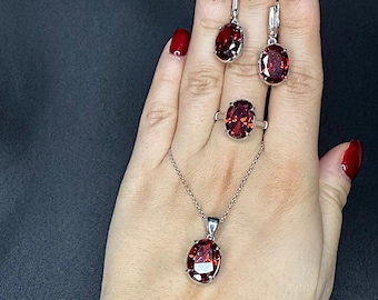 May include: A jewellery set comprising a silver necklace, ring, and earrings, each with a large, oval, deep red gemstone. The earrings are drop style, and the ring and pendant have a simple design. The set is displayed on a hand with red nail polish.