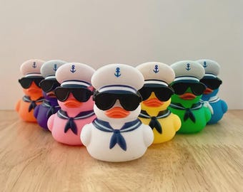 May include: A collection of seven rubber ducks arranged in a semi-circle. Each duck is wearing a white sailor hat with a blue anchor emblem and black sunglasses. The ducks are colored white, pink, yellow, green, blue, purple, and orange.