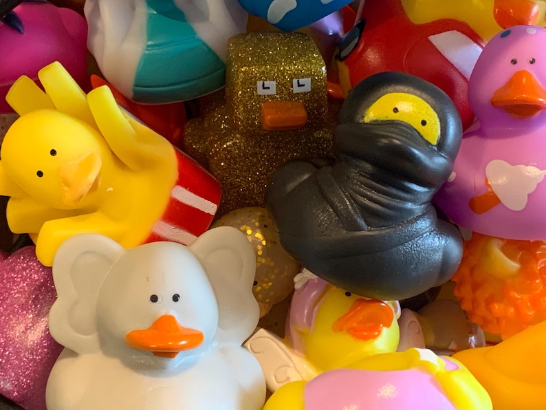 May include: A collection of colorful rubber duck toys, including a yellow duck with a popcorn container, a white duck with elephant ears, a black duck with a ninja mask, a purple duck with a crown, and a gold duck with glitter.
