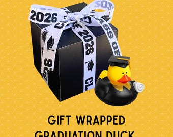 May include: A black gift box tied with a white ribbon printed with "2026" and graduation caps. A yellow rubber duck wearing a graduation cap and gown is next to the box. The text "GIFT WRAPPED GRADUATION DUCK" is below the box.