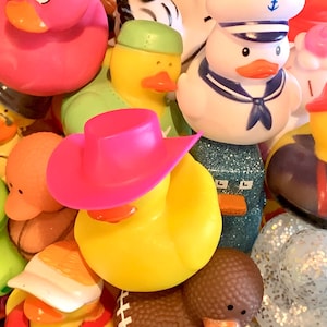 May include: A collection of colorful rubber duck toys, including a pink duck with a cowboy hat, a white duck with a sailor hat, a green duck with a green hat, and a yellow duck with a red hat. The ducks are arranged in a pile, with some of them overlapping.