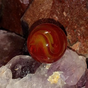 Könnte beinhalten: Eine rote und gelbe Glaskugel mit wirbelnden Mustern liegt auf einem Haufen violetter und klarer Kristalle.