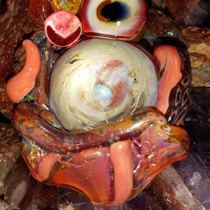 May include: A glass sculpture with a swirling, iridescent orb, surrounded by colorful, textured glass pieces. The sculpture features a large eye with a black pupil and a yellow iris, and a smaller eye with a blue pupil and a white iris.