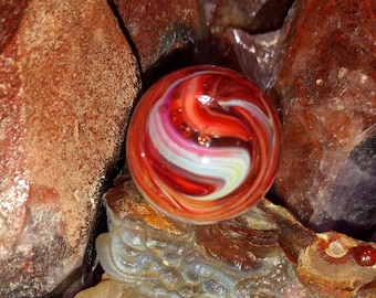 May include: A red, white, and pink swirl glass marble resting on a bed of brown, red, and purple stones.