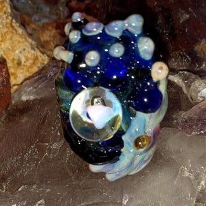 May include: A blue glass bead with white and gold accents. The bead has a round, clear glass center with a pink swirl inside.