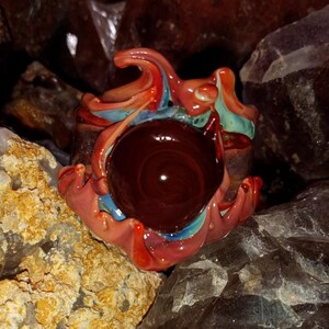 May include: A glass art piece with a red, orange, and blue swirl design. The piece has a large, dark red center and is surrounded by a variety of crystals.