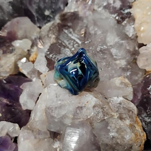 May include: A handcrafted glass ring with a unique, abstract design. The ring features swirls of blue, white, and black colors. The ring is set against a background of various crystals and stones.