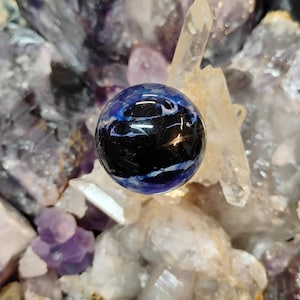 May include: A polished, spherical gemstone with a dark center and swirling blue and white patterns. The orb is surrounded by a collection of raw quartz crystals and amethyst clusters, creating a natural, earthy aesthetic.