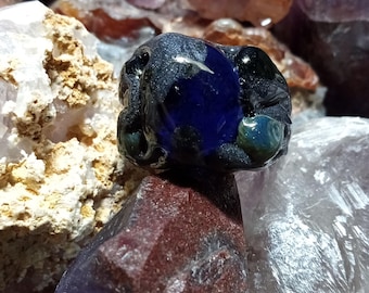 Peut inclure: Une perle de verre bleu foncé avec des tourbillons de vert et de noir, entourée de divers cristaux.