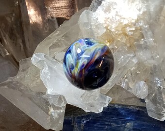 Peut inclure: Un marbre en verre bleu avec des tourbillons blancs et jaunes. Le marbre est posé sur un lit de cristaux de quartz transparents.