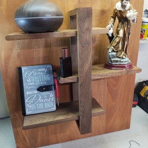 May include: A wooden shelf unit with three shelves. The top shelf has a dark brown diffuser, the middle shelf has a bottle of liquid and a red tool, and the bottom shelf has a black journal with the text "Always be waiting for the storm to pass... Learning to Dance in the Rain". A small statue of a person in a brown robe is on the right side of the shelf unit.
