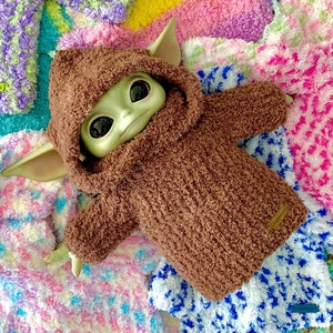 A Baby Yoda doll wearing a brown Snuggle Hoodie laying on a bed of multicolored Snuggle Hoodies