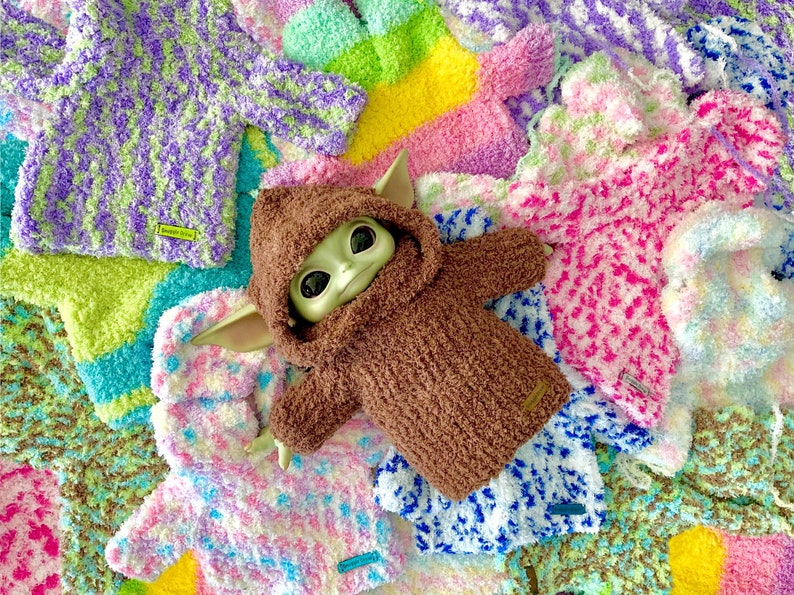 A Baby Yoda doll wearing a brown Snuggle Hoodie laying on a bed of multicolored Snuggle Hoodies