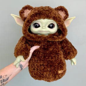 Puede incluir: Muñeco de peluche de un personaje de una popular franquicia de ciencia ficción, que lleva un traje con capucha marrón con orejas de oso. El muñeco tiene grandes ojos negros, piel verde y orejas puntiagudas. Una pequeña etiqueta rectangular es visible en el traje.
