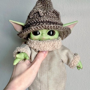 May include: A green plush toy wearing a brown crocheted hat and a beige coat. The toy has large eyes and a small mouth.