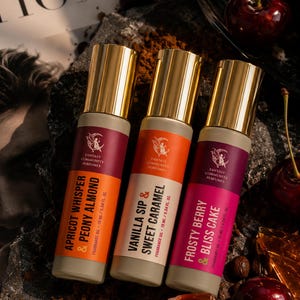 May include: Three perfume bottles with gold caps and labels that read "Apricot Whisper & Peony Almond", "Vanilla Sip & Sweet Caramel", and "Frosty Berry & Bliss Cake". The bottles are on a dark, textured surface with cherries.