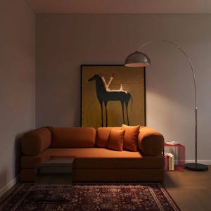以下が含まれることがあります： 2つのボルスタークッションと2つのスクエアクッションを備えた錆色のソファ。黒い馬が描かれた額縁入りの絵画の下に配置されています。右側には金属製のドーム型シェードが付いた背の高いアーチ型のフロアランプがあり、その隣には赤いワイヤー製のサイドテーブルが置かれています。