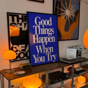 May include: A room with a wooden and metal shelf displaying art prints and decorative items. A blue poster with white text that reads "Good Things Happen When You Try" is the focal point. Other art pieces and orange globe lamps add to the aesthetic.