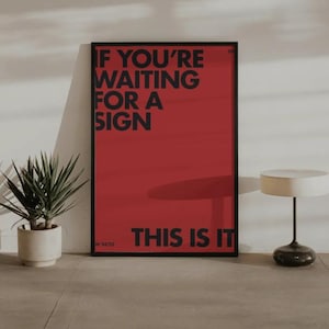 May include: A framed red poster with black text that reads "IF YOU'RE WAITING FOR A SIGN THIS IS IT." A potted plant and a small table lamp are in the foreground. The poster is leaning against a wall.