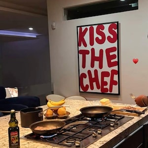 Könnte beinhalten: Ein rechteckiges Wandbild mit dem fetten roten Schriftzug KISS THE CHEF auf weißem Hintergrund, schwarz gerahmt. Das Kunstwerk hängt über einer Kücheninsel mit einem Gasherd, Kochpfannen, einer Flasche Olivenöl und verschiedenen Früchten.