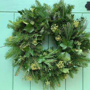 May include: A circular wreath constructed from fresh evergreen boughs, including pine needles, spruce branches, and glossy green ivy leaves. The arrangement is accented with clusters of small, pale yellow-green flower buds distributed throughout the foliage.