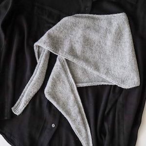 Pode incluir: Um cachecol triangular de malha cinza-claro com uma aparência texturizada e mesclada, com pontas longas e afiladas. O cachecol está sobreposto a uma camisa preta de manga comprida com botões, destacando o contraste entre a malha cinza e o tecido escuro.
