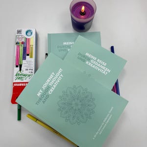 Könnte beinhalten: Eine Auswahl an Journalen und Stiften für kreative Zwecke. Die Journale sind hellgrün mit dem Text "MY JOURNEY THROUGH THOUGHT AND CREATIVITY" und einem Mandala-Design. Eine brennende lila Kerze und eine Box mit Stiften sind ebenfalls zu sehen.