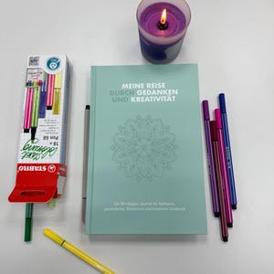 Könnte beinhalten: Ein türkisfarbenes Journal mit dem deutschen Text "MEINE REISE DURCH GEDANKEN UND KREATIVITÄT" und einem Mandala-Design, umgeben von bunten Stiften, einem gelben Textmarker, einer Packung Stabilo-Stiften und einer brennenden lila Kerze.